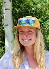 Person wearing a yellow cap with a scenic design, standing outdoors with greenery in the background