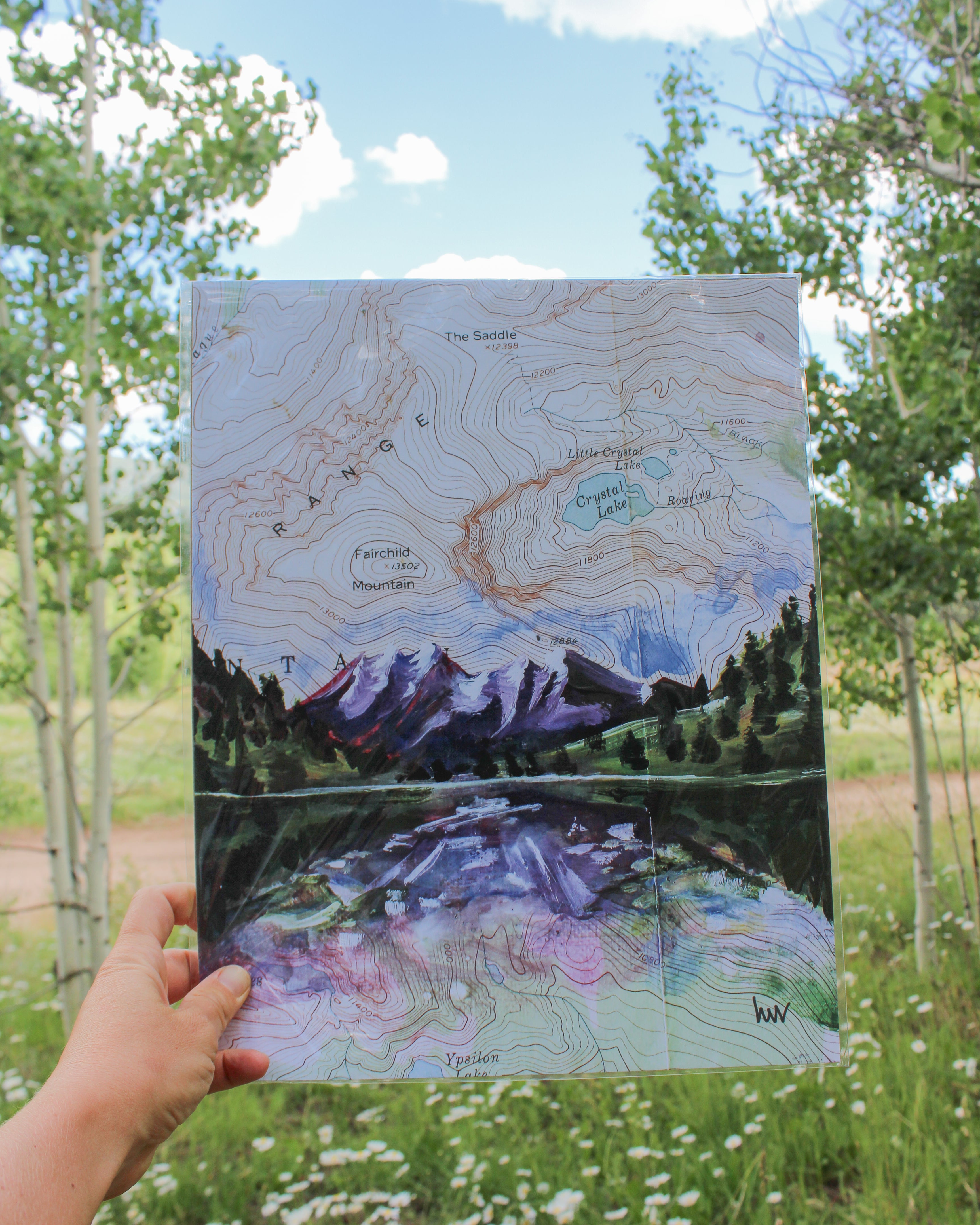 Hand holding a map of a mountain range with a lake, surrounded by trees and grass.