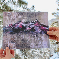 A high-quality print artwork featuring a stylized mountain landscape, held up by a pair of hands.
