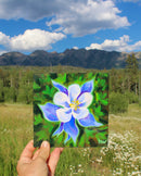 Hand holding a painting of a blue and white flower against a natural background with mountains and sky.