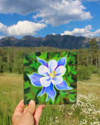 Hand holding a painting of a blue and white flower against a natural background with mountains and sky.