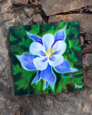 Painting of a blue flower on a small canvas with a rocky background