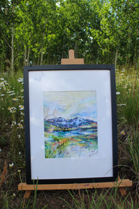 Framed artwork of a landscape with mountains and water on an easel outdoors.