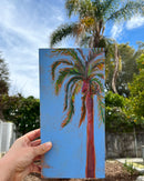 Hand holding a painting of a palm tree against a blue sky with trees in the background