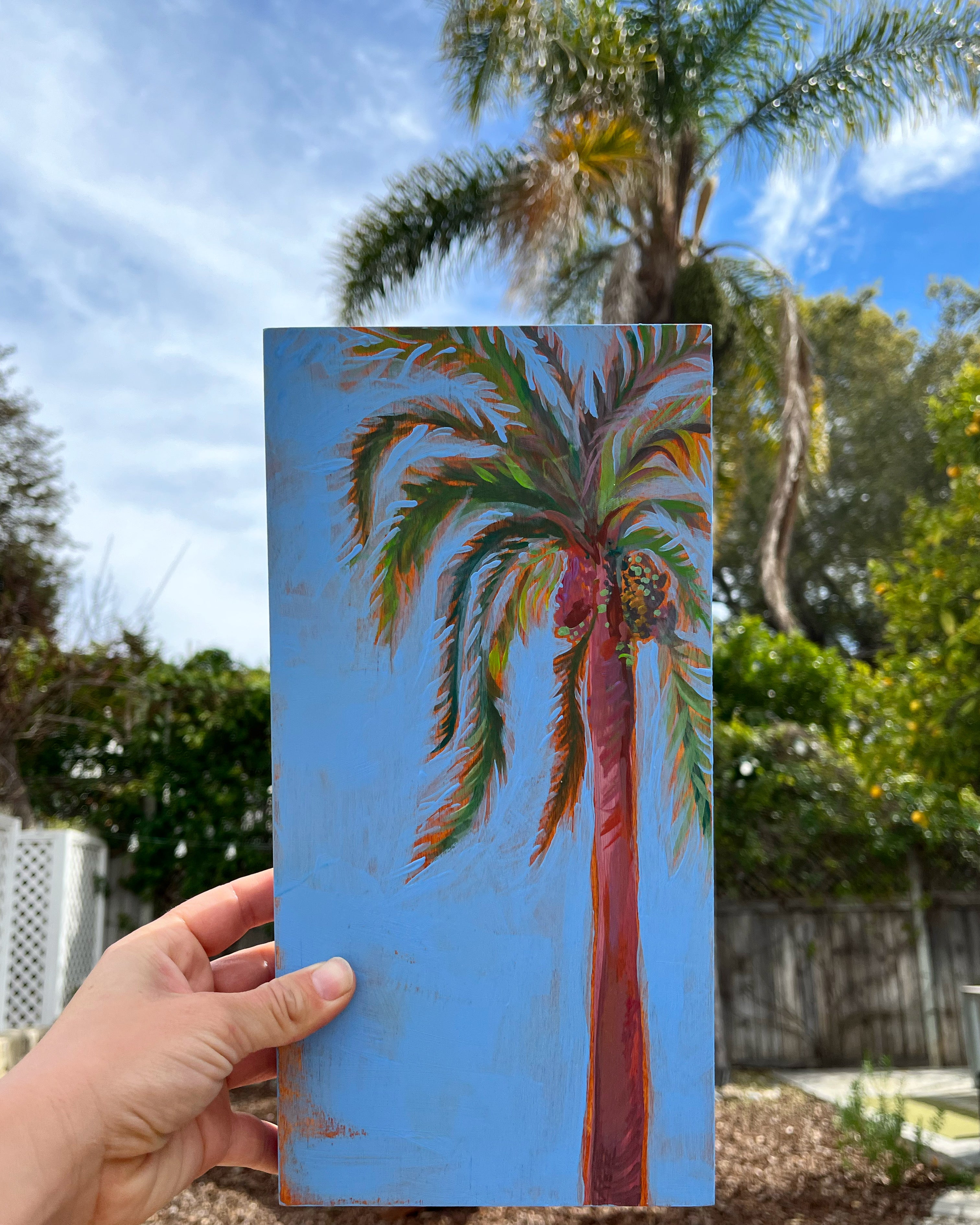 Hand holding a painting of a palm tree against a blue sky with trees in the background