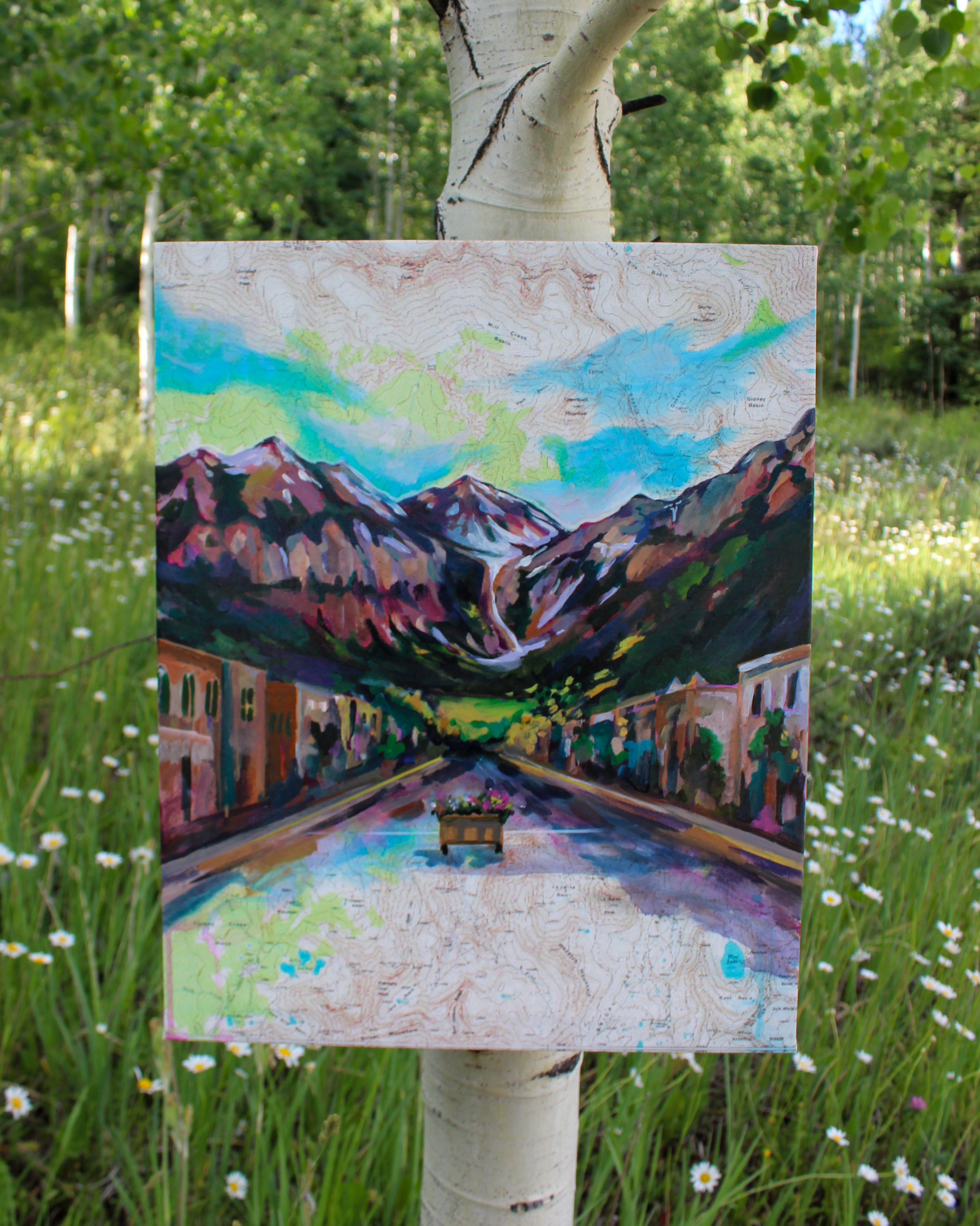 Painting of a mountain village scene on an easel in a natural setting with trees and grass.