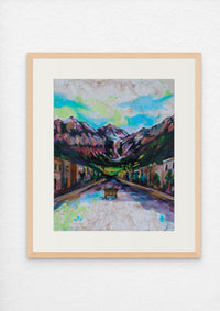 Framed painting of a mountainous landscape with a town below on a white wall.