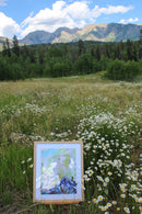 Painting of a mountain scene on an easel in a field with wildflowers and mountains in the background