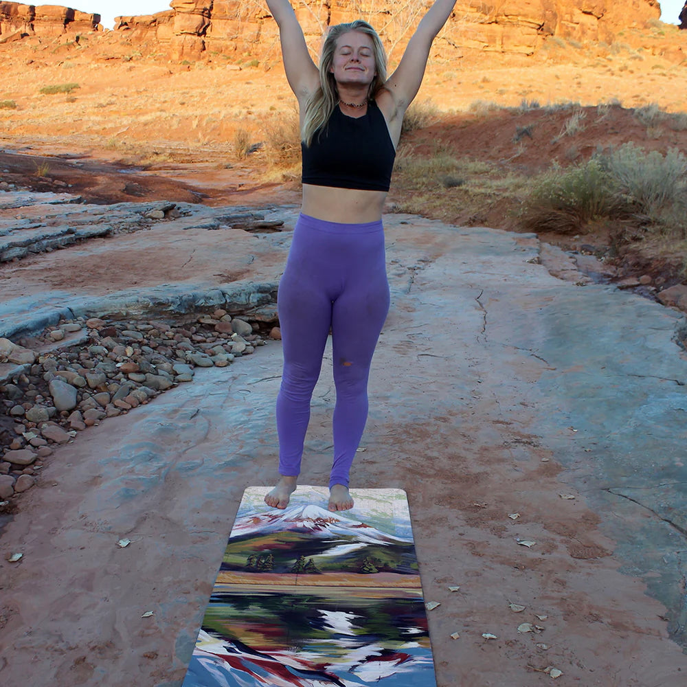 Woman in purple leggings and black top practicing yoga on a colorful mat in a desert landscape.