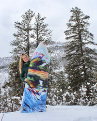 Woman holding a yoga mat with a mountain painting on it up while standing in the snow.