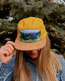 Person wearing a yellow cap with a scenic design, standing in front of trees.